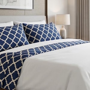 Puede incluir: Una cama con funda nórdica blanca y un camino de mesa azul marino con un patrón de cuatrifolio blanco. La cama incluye dos almohadas a juego con patrón de cuatrifolio azul marino y dos almohadas blancas lisas, frente a un cabecero gris claro.