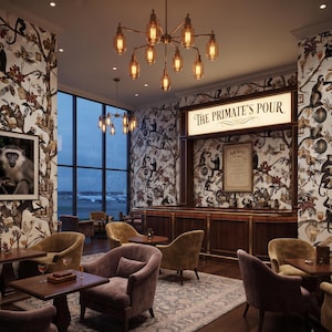 May include: A bar interior named The Primate's Pour featuring wallpaper with monkey illustrations, dark wood paneling, a large illuminated sign, amber glass chandeliers, and several upholstered armchairs arranged around wooden tables.