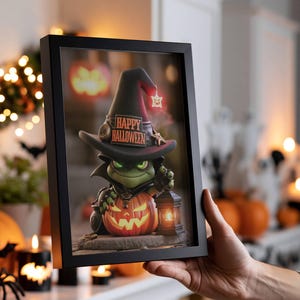 May include: A black framed print featuring a green creature wearing a pointed witch hat with the text HAPPY HALLOWEEN. The creature sits behind a glowing orange jack-o'-lantern and holds a small black lantern, set against a dark background.