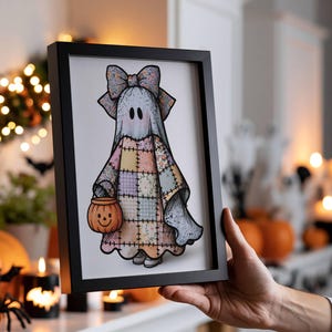 May include: A framed art print featuring a cartoon ghost wearing a large patterned bow and a patchwork quilt-style sheet in pastel colours. The ghost holds a small orange jack-o'-lantern bucket. The illustration has a black outline.