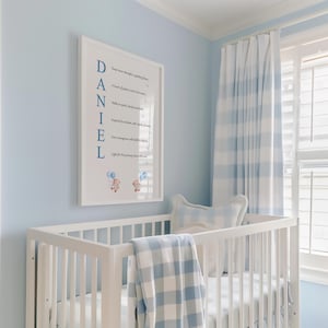 May include: A white crib with a blue and white checkered blanket and pillow. A framed print on the wall reads "DANIEL" with inspirational text. The room has light blue walls and matching curtains.
