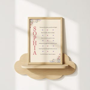 以下が含まれることがあります： 左側にピンクの大文字で「SOPHIA」と書かれた額入りのプリント。プリントはベージュの背景に花のアクセントとインスピレーションを与えるテキストがあります。木製の雲型の棚の上に置かれています。