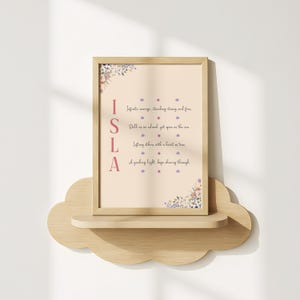 以下が含まれることがあります： ピンク色の「ISLA」という名前と詩が書かれた額入りのプリント。隅には花の装飾が施されています。プリントは木製の雲型の棚に置かれています。背景は薄いベージュ色です。