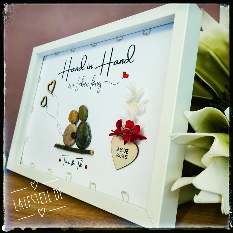 May include: A white framed wall art piece featuring pebble art of two figures sitting on a branch, holding two heart-shaped balloons. The text reads Hand in Hand ein Leben lang, Tina & Tobi, and 25.07.2025 on a wooden heart with red flowers.