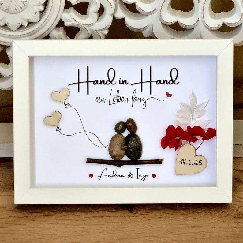 May include: A white framed wall art piece featuring pebble art of two figures sitting on a twig. The text reads Hand in Hand ein Leben lang and Andrea & Ing. with a heart shape displaying 14.6.25. Includes two wooden heart balloons and red floral accents.