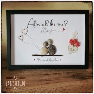May include: A framed pebble art piece featuring two figures made of stones sitting on a twig. The text reads After all this time? Always, Vanessa & Maximilian, and 06.12.2025. It includes two wooden heart balloons, red floral accents, and a heart-shaped tag.