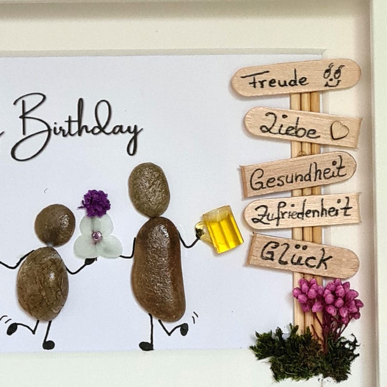 May include: A framed birthday art piece featuring pebble figures holding a flower and a beer mug. Wooden signs point to 'Freude', 'Liebe', 'Gesundheit', 'Zufriedenheit', and 'Gl&uuml;ck'. The artwork includes decorative flowers and moss.