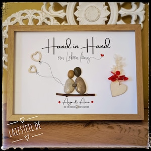 May include: A framed wall art piece featuring two pebble figures sitting on a twig, with two heart-shaped balloons drawn in black ink. The text reads Hand in Hand ein Leben lang, Anja & Arne, and the dates 02.10.2000 and 02.10.2025 with a small heart.