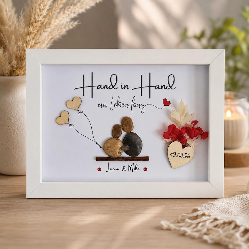 May include: A white framed wall art piece featuring pebble art of two figures sitting on a branch. The text reads Hand in Hand ein Leben lang, Lena & Mike, and 19.09.26. It includes two wooden heart balloons, red dried flowers, and a wooden heart.