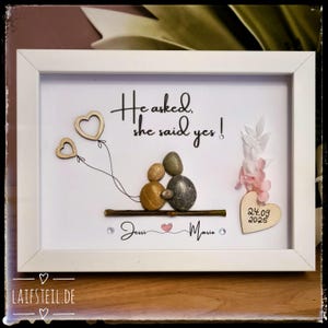 May include: A white framed wall art piece featuring pebble art of two figures sitting on a twig. The text reads He asked, she said yes! and Jessi and Mario with a small heart. A wooden heart displays the date 24.09.2025 with pink floral accents.