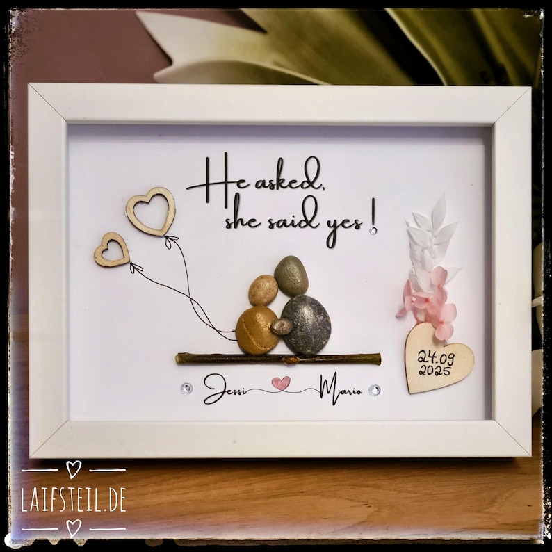 May include: A white framed wall art piece featuring pebble art of two figures sitting on a twig. The text reads He asked, she said yes! and Jessi and Mario with a small heart. A wooden heart displays the date 24.09.2025 with pink floral accents.
