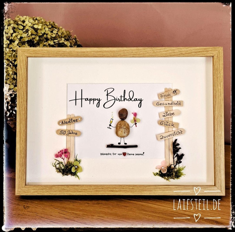 May include: A framed birthday art piece with the words "Happy Birthday" in elegant script. The artwork features a pebble figure holding a champagne glass and flowers, with wooden signs pointing to well wishes. The frame is light brown.