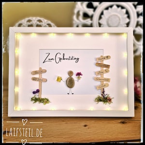 May include: A framed birthday art piece with a white frame and warm white LED lights. The artwork features a pebble figure holding a beer and flowers, with wooden signs and the text "Zum Geburtstag".