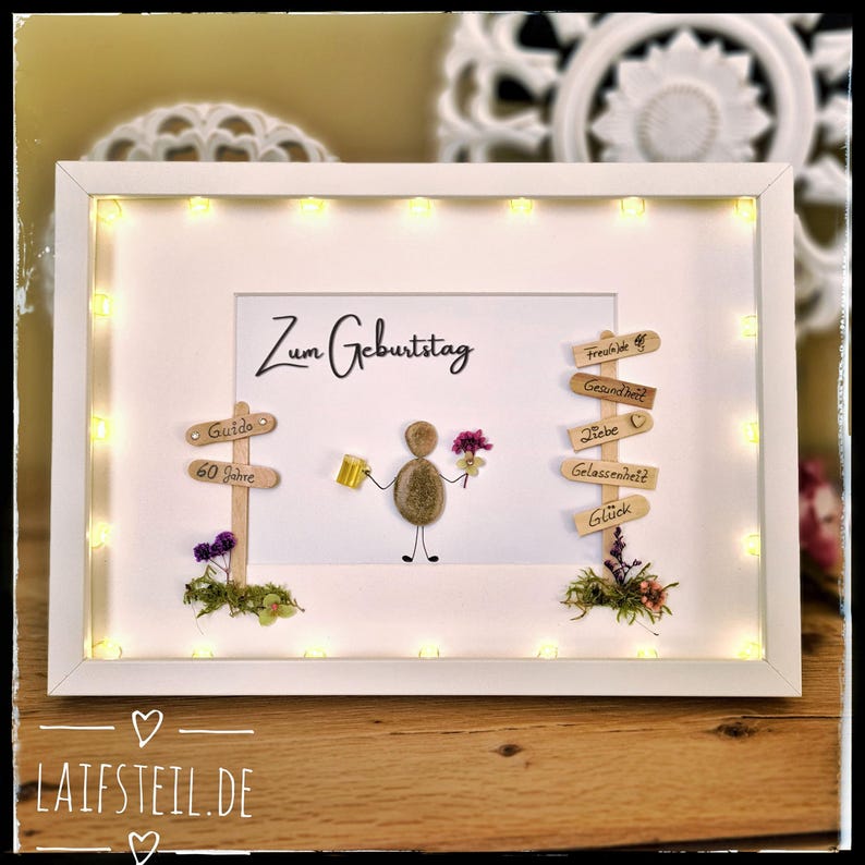 May include: A framed birthday art piece with a white frame and warm white LED lights. The artwork features a pebble figure holding a beer and flowers, with wooden signs and the text "Zum Geburtstag".