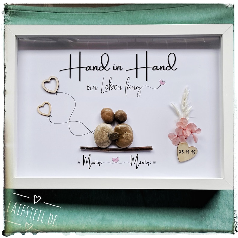 May include: A white framed wall art piece featuring pebble art of two figures sitting on a twig. The text reads Hand in Hand ein Leben lang, Matzi, and Mietzi. Includes two wooden heart outlines, dried pink flowers, and a heart with 28.11.15.
