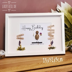 May include: A framed artwork with a white border. The artwork features a pebble person holding a champagne glass and flowers, with the words "Happy Birthday" above. Wooden signs and dried flowers are also included. The frame measures 9x12.6x1.2 inches.