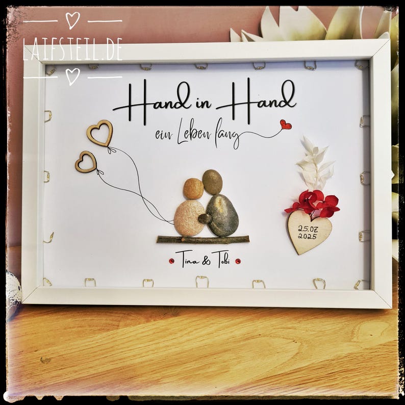 May include: A white framed wall art piece featuring pebble art of two figures sitting on a twig, holding two wooden heart balloons. The text reads Hand in Hand ein Leben lang, Tina & Tobi, and 25.07.2025 on a wooden heart with red flowers.
