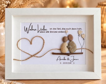 May include: A white framed wall art piece featuring pebble art of two figures sitting on a branch. The text reads "Wahre Liebe ist das Seil, das auch dann hält, wenn alle Stricke reißen", "Annika & Jonas", and "23.10.2026". A twine heart is on the left.