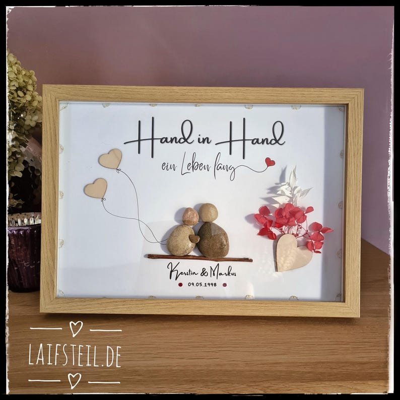 May include: A framed pebble art piece featuring two figures sitting on a branch, with the text "Hand in Hand ein Leben lang", "Kerstin & Markus", and "09.05.1998". The design includes two wooden heart balloons, red dried flowers, and a wooden heart.
