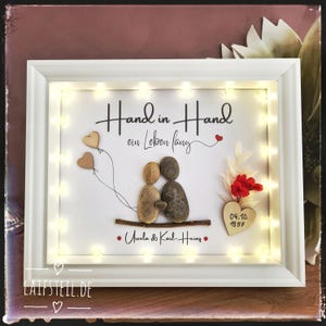 May include: A white shadow box frame containing pebble art of two figures sitting on a branch, with the text Hand in Hand ein Leben lang, Ursula & Karl-Heinz, and the date 04.10.1957. The display features warm LED lights, two wooden heart balloons, and red dried flowers.