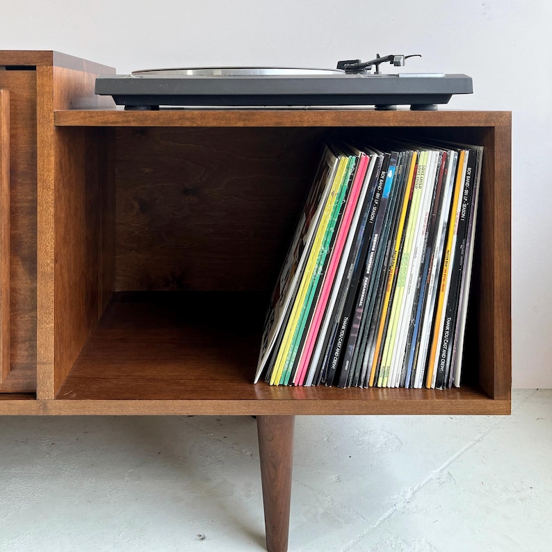 May include: A wooden record player stand with a turntable on top and a shelf below for storing vinyl records. The shelf is filled with a variety of records.