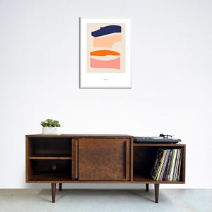 May include: A mid-century modern style wooden entertainment center with a record player, shelves, and a cabinet door. The entertainment center is in front of a white wall with a framed abstract art print featuring geometric shapes in blue, orange, and pink.