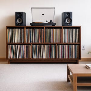 May include: A wooden record cabinet filled with vinyl records. Two black speakers and a turntable sit on top. The cabinet is in a living room with a light gray sofa, a wooden coffee table, and a white rug.
