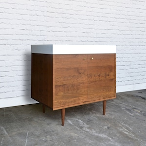 May include: A mid-century modern bathroom vanity with two doors and a white countertop. The vanity is made of walnut wood and has brass hardware. The vanity is standing on four tapered legs.