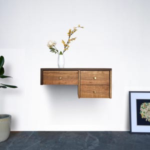 May include: A floating wall shelf with two drawers made of light brown wood with brass knobs. The shelf is mounted on a white wall and has a vase with yellow flowers on top.