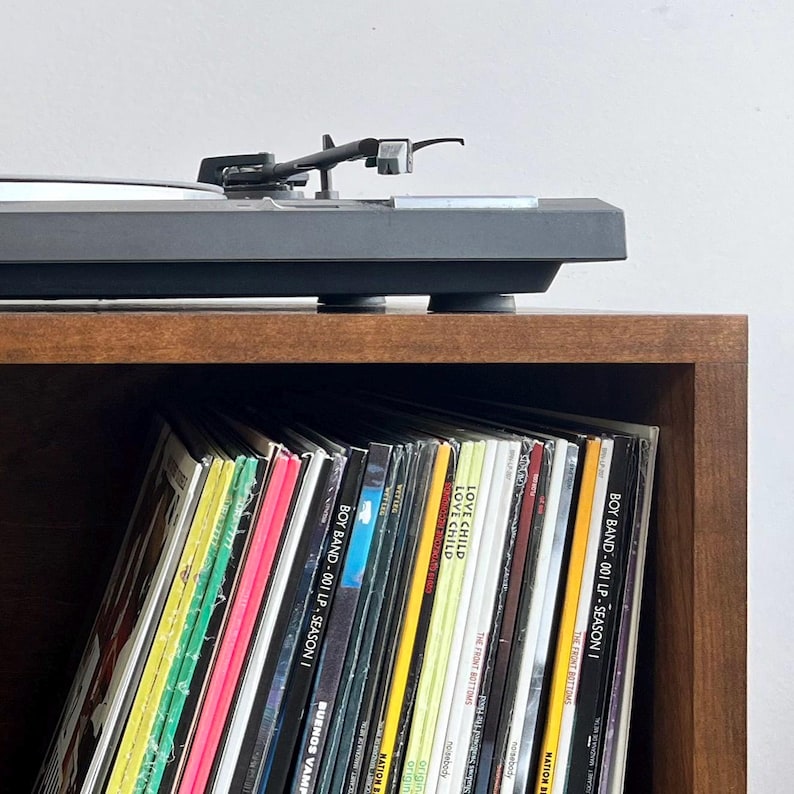 May include: A wooden shelf unit with a turntable on top and a collection of vinyl records on the lower shelf. The records are organized vertically and include titles like "BOY BAND - 001 LP - SEASON I" and "LOVE CHILD".