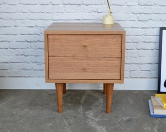 May include: A two-drawer wooden nightstand with brass knobs and tapered legs. The nightstand is made of light wood and has a simple, modern design.