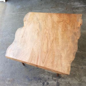 May include: A large, natural wood coffee table with a live edge top and a light brown finish. The table has a unique, organic shape and is supported by two simple legs.