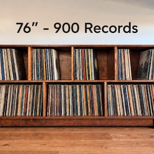 May include: A dark brown wooden storage unit with five rectangular compartments filled with vinyl records. The unit measures 76 inches in width and has a capacity for 900 records, as indicated by the text 76" - 900 Records above the unit.
