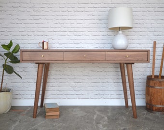 May include: A mid-century modern walnut desk with three drawers and brass hardware. The desk has tapered legs and is sitting on a concrete floor. A lamp with a white shade is on the right side of the desk.