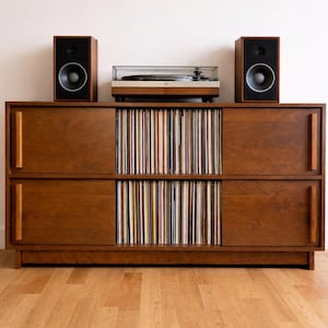 May include: A wooden record cabinet with a turntable and two speakers on top. The cabinet is a warm brown color and has two sets of sliding doors. The cabinet is filled with vinyl records.