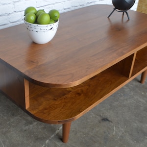 May include: A mid-century modern coffee table with a rounded oval top and two open shelves. The table is made of dark wood and has a natural finish. The table is sitting on a concrete floor.