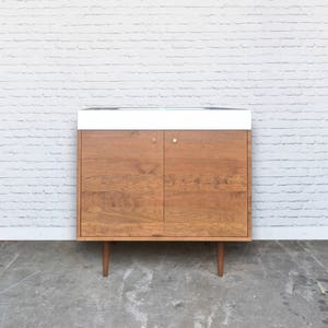 May include: A mid-century modern style cabinet with two doors and a white countertop. The cabinet is made of wood with a light brown finish and has brass hardware. The cabinet is sitting on a concrete floor in front of a white brick wall.
