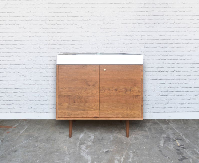 May include: A mid-century modern style cabinet with two doors and a white countertop. The cabinet is made of wood with a light brown finish and has brass hardware. The cabinet is sitting on a concrete floor in front of a white brick wall.