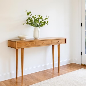May include: A mid-century modern wooden console table with three drawers and tapered legs. A white vase with green foliage and a small white bowl sit on top. The table is made of light-colored wood.