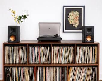 May include: A dark wood media console with eight open storage compartments filled with vinyl records. A black turntable with a clear dust cover sits on top, flanked by two black speakers with copper-colored woofers and a vase of yellow flowers.