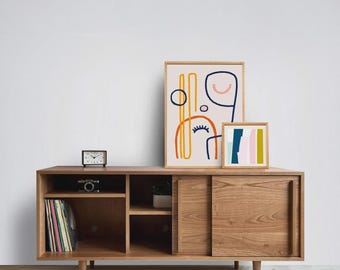 May include: A wooden media console with sliding doors, open shelving, and tapered legs. On top sit two framed prints featuring abstract line art and color blocks. A small analog clock and a camera are placed on the open shelves.
