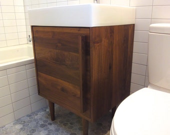 May include: A white porcelain bathroom sink with a chrome faucet sits atop a dark wood cabinet with a single door. The cabinet has four legs and is set on a gray hexagonal tile floor. A white toilet is visible in the background.