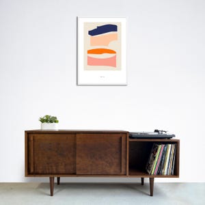 May include: A mid-century modern style wooden cabinet with sliding doors and a record player on top. The cabinet has a shelf on the right side that is filled with vinyl records. A framed abstract print with geometric shapes in blue, orange, and pink hangs on the wall above the cabinet.