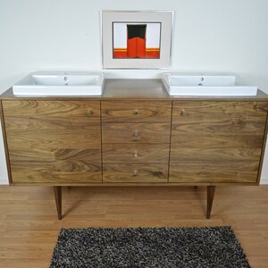 Pu&ograve; includere: Un mobile da bagno in legno di noce con due lavabi in porcellana bianca e quattro cassetti. Il mobile ha un design moderno di met&agrave; secolo con gambe affusolate.