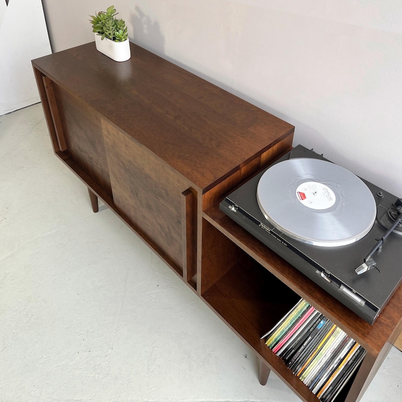 May include: A dark brown wooden record player stand with sliding doors and open shelves. The stand has a turntable on top and a collection of vinyl records on the bottom shelf.