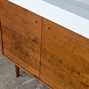 May include: A wooden bathroom vanity with two cabinet doors and a white countertop. The cabinet doors have brass knobs and the wood has a natural grain pattern.