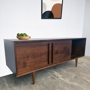 May include: A dark brown wooden media console with two sliding doors and a shelf. The console has a modern design with tapered legs and is sitting on a concrete floor.