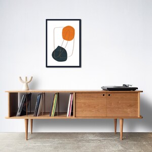 May include: A mid-century modern style record player sits on top of a wooden cabinet with open shelves. The cabinet has a sliding door on the right side. The shelves are filled with vinyl records. A framed print with abstract shapes in orange and blue hangs on the wall above the cabinet.