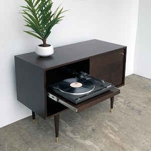 May include: A dark brown wooden record player stand with a sliding drawer that reveals a turntable and a record. The stand has gold-colored legs and a small door on the right side.