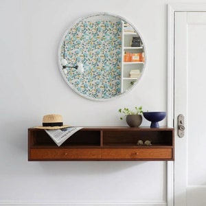May include: A wooden floating shelf with two drawers and a round mirror with a floral pattern. The shelf is mounted on a white wall and has a hat, newspaper, and two small decorative objects on top.